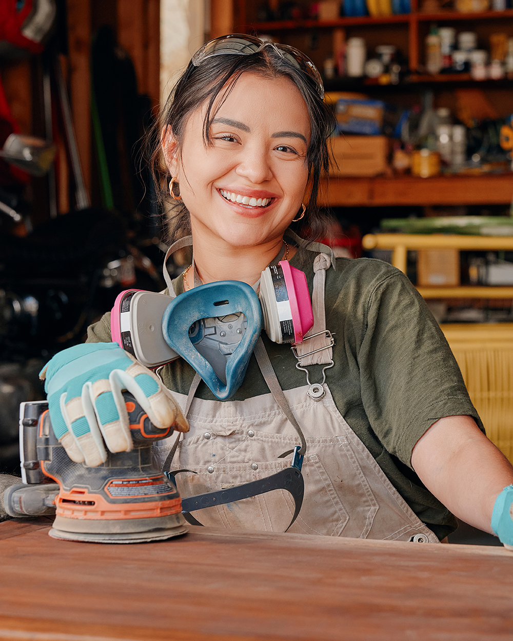 Woman smiling in a workshop while sanding a wooden tabletop with protective gloves, goggles, and sander