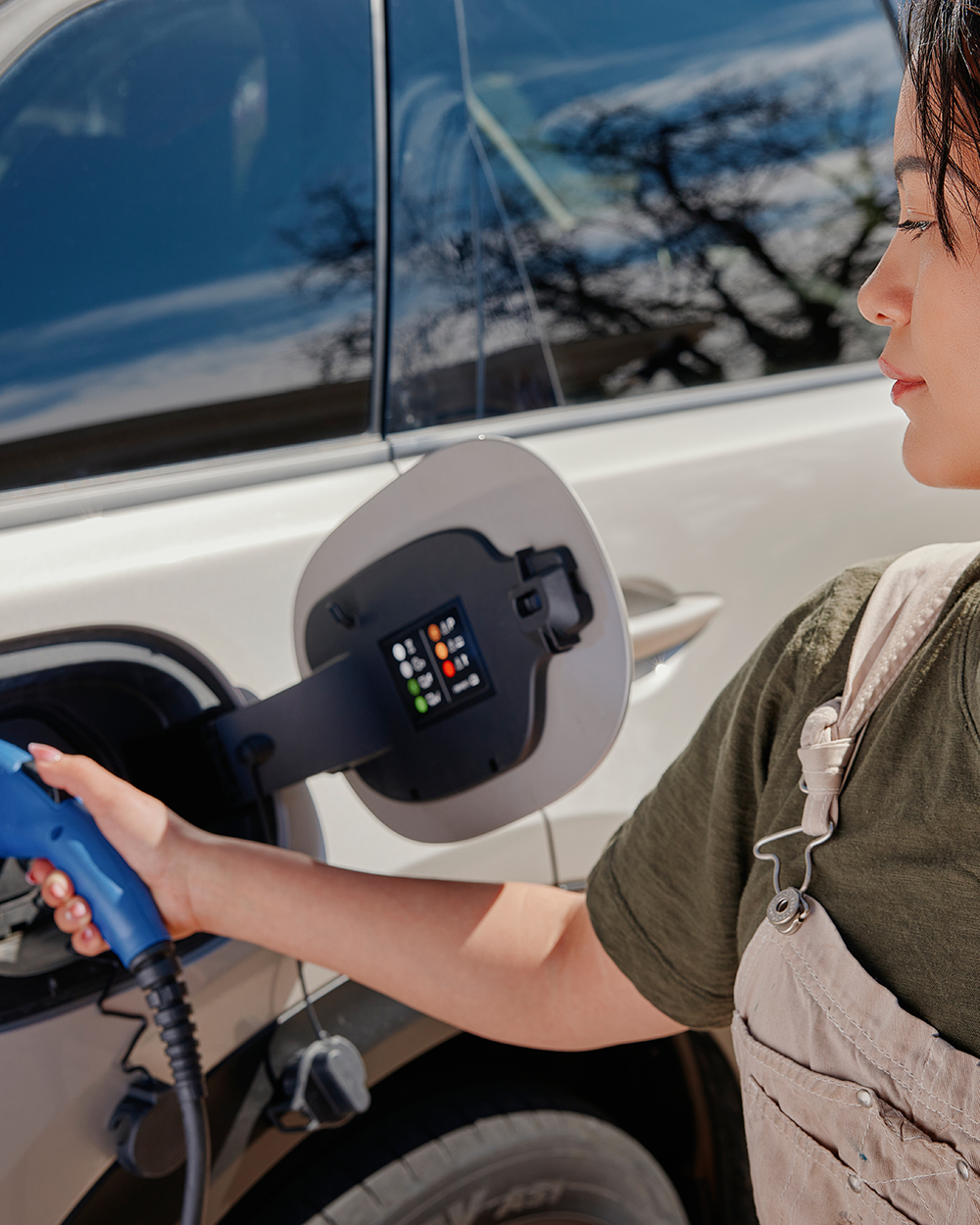 Woman wearing overalls plugs in an electric vehicle to charge during the day