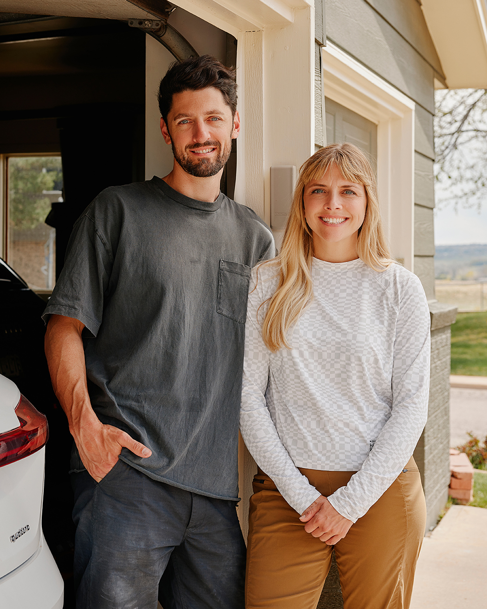 Man and woman smiling standing outside their garage next to a white EV with a Colorado license plate