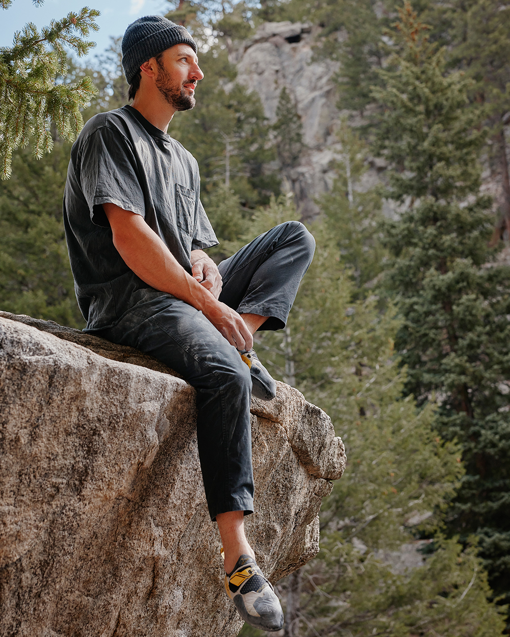 Man sitting on the edge of a large rock, looking out at a forested landscape in climbing shoes