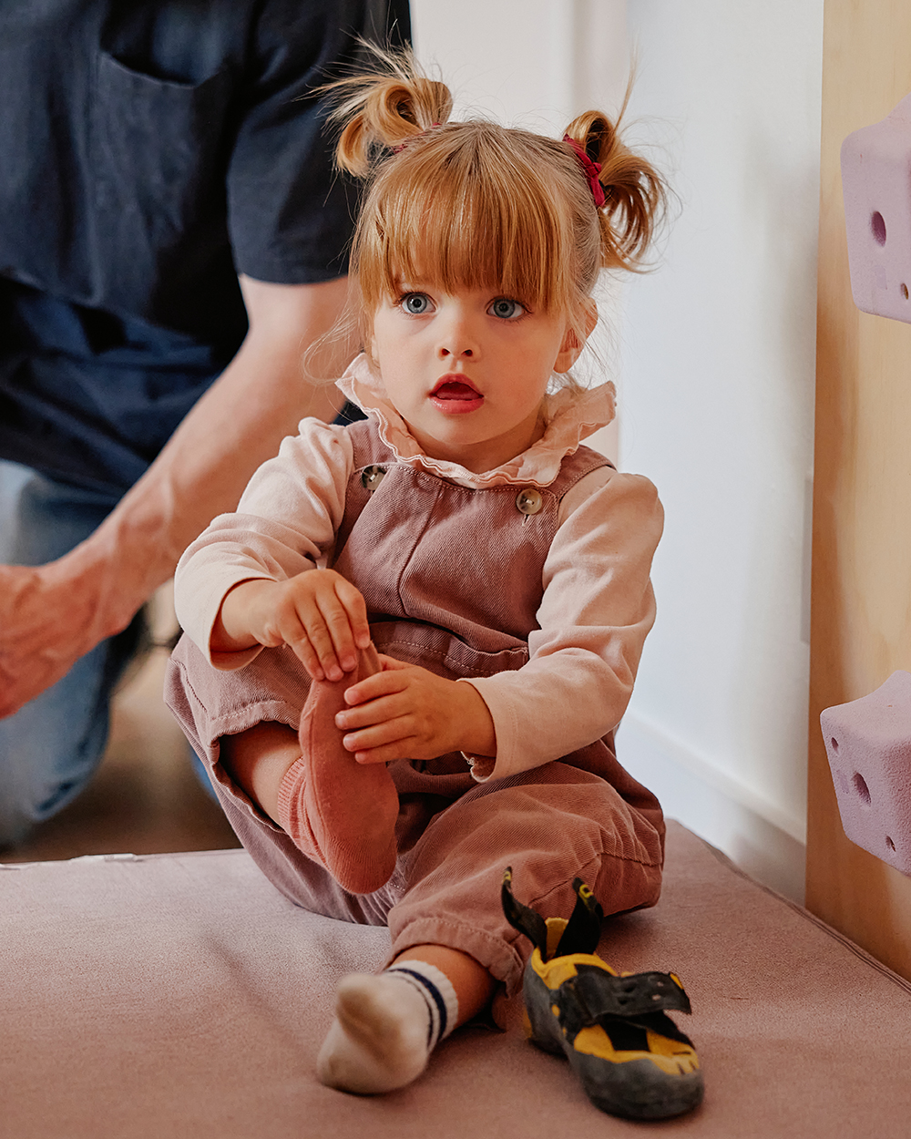 Young girl sitting beside a climbing wall, putting on climbing shoes with help from an adult