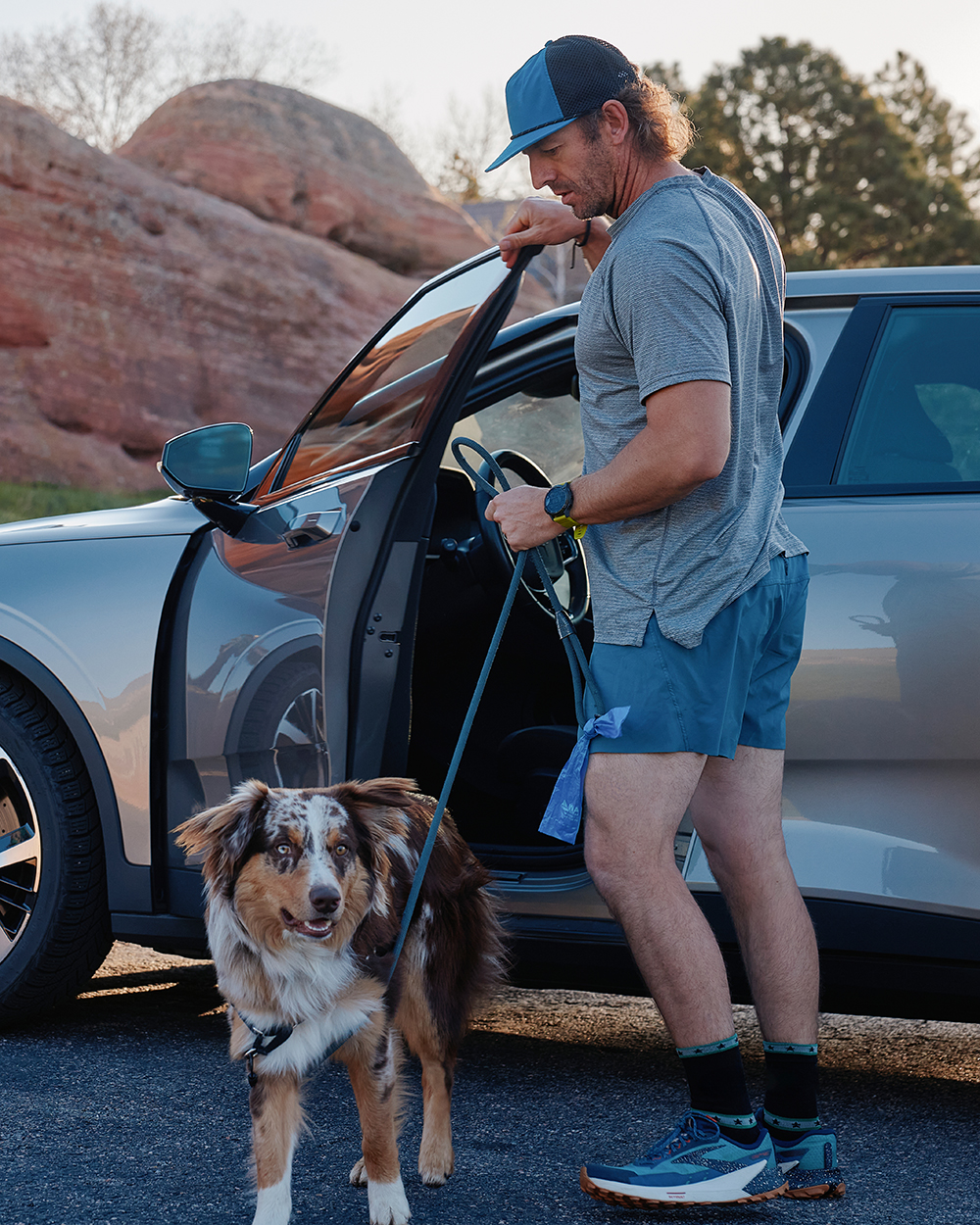 Man holding a leash while standing next to an electric vehicle with his dog in a scenic outdoor parking area