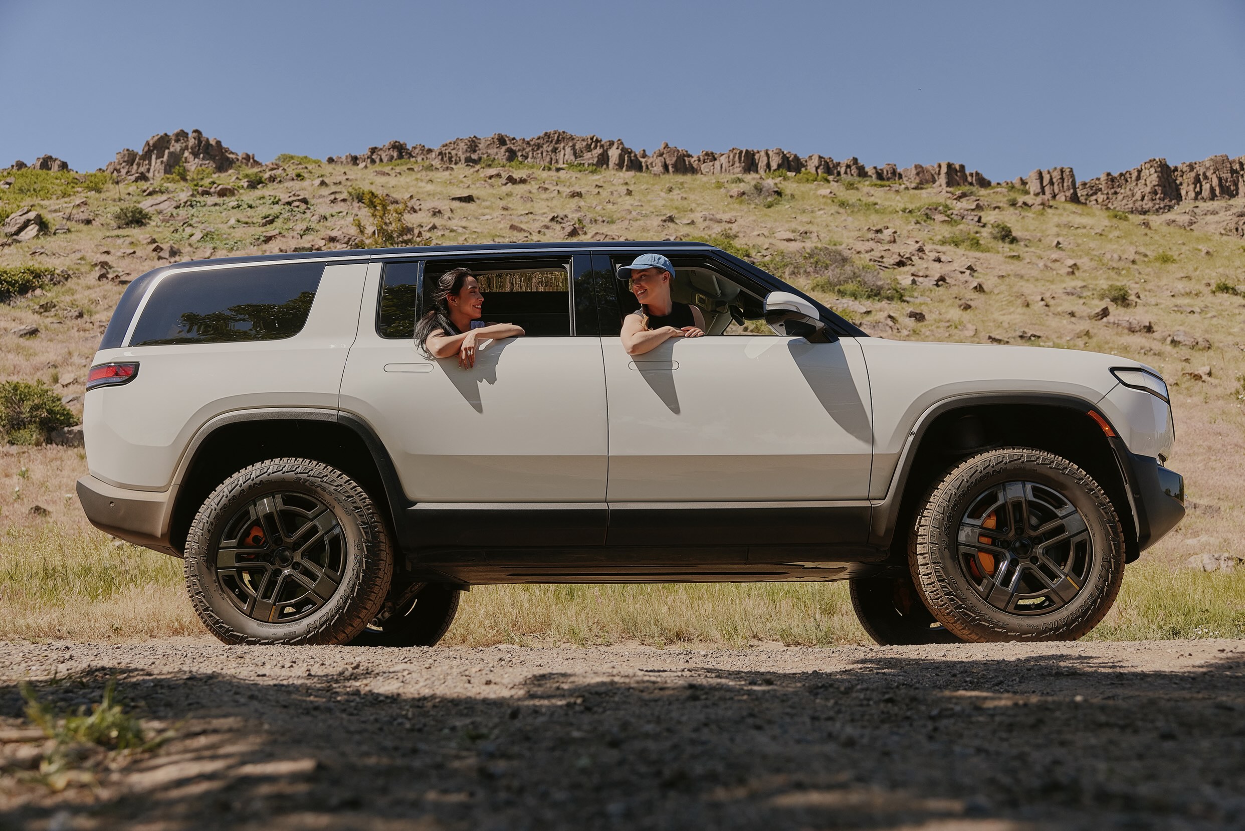 Two people stick their heads out the windows of a SUV on a dirt road with rocky terrain in the background.