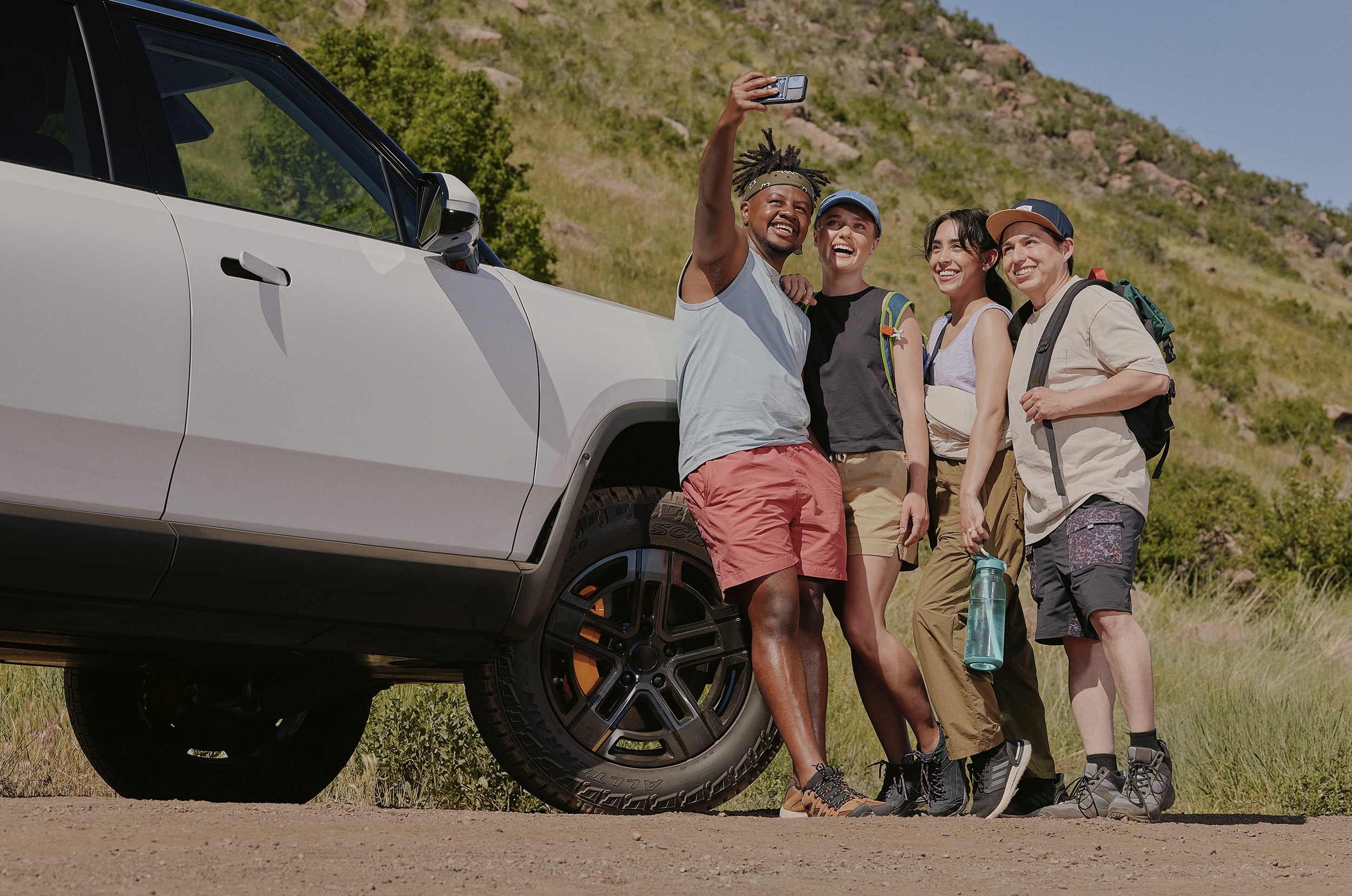 Four people take a selfie in front of an SUV on a dirt road.