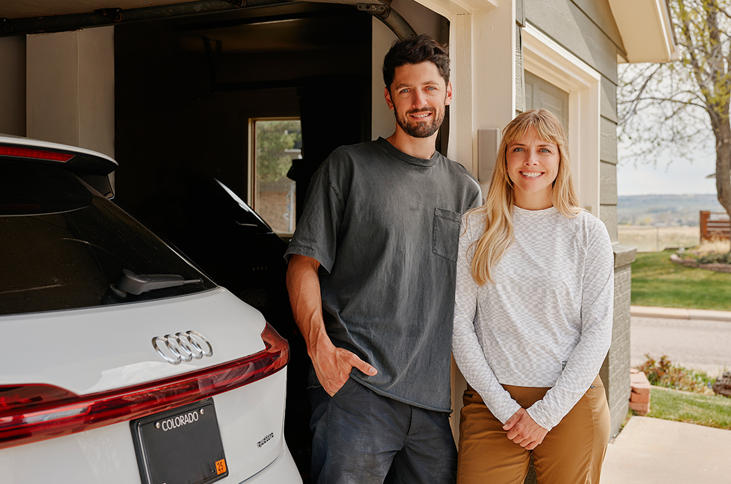 Man and woman smiling standing outside their garage next to a white EV with a Colorado license plate