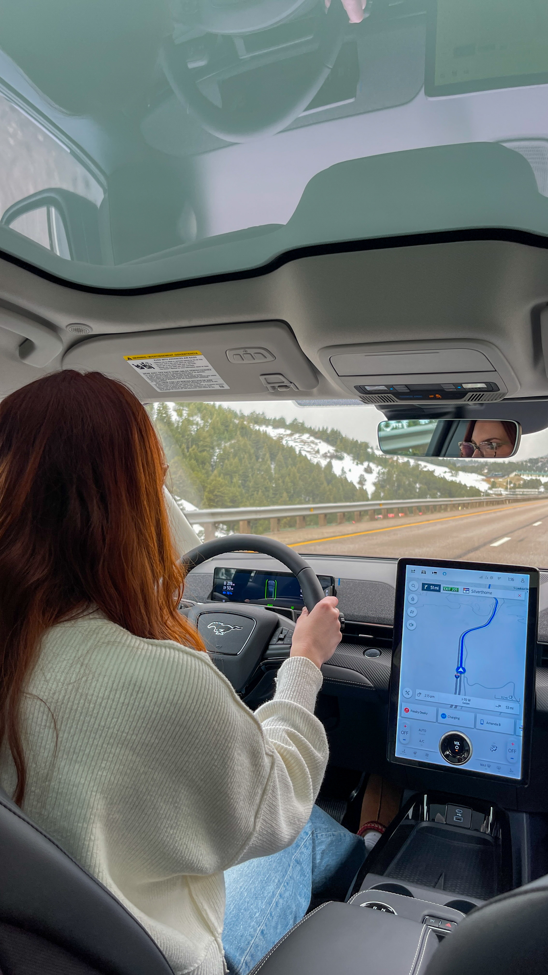 Amanda Bittner driving her Ford Mustang Mach-E EV, viewed from the rear-right passenger seat. The touchscreen status console is visible, showing GPS navigation.