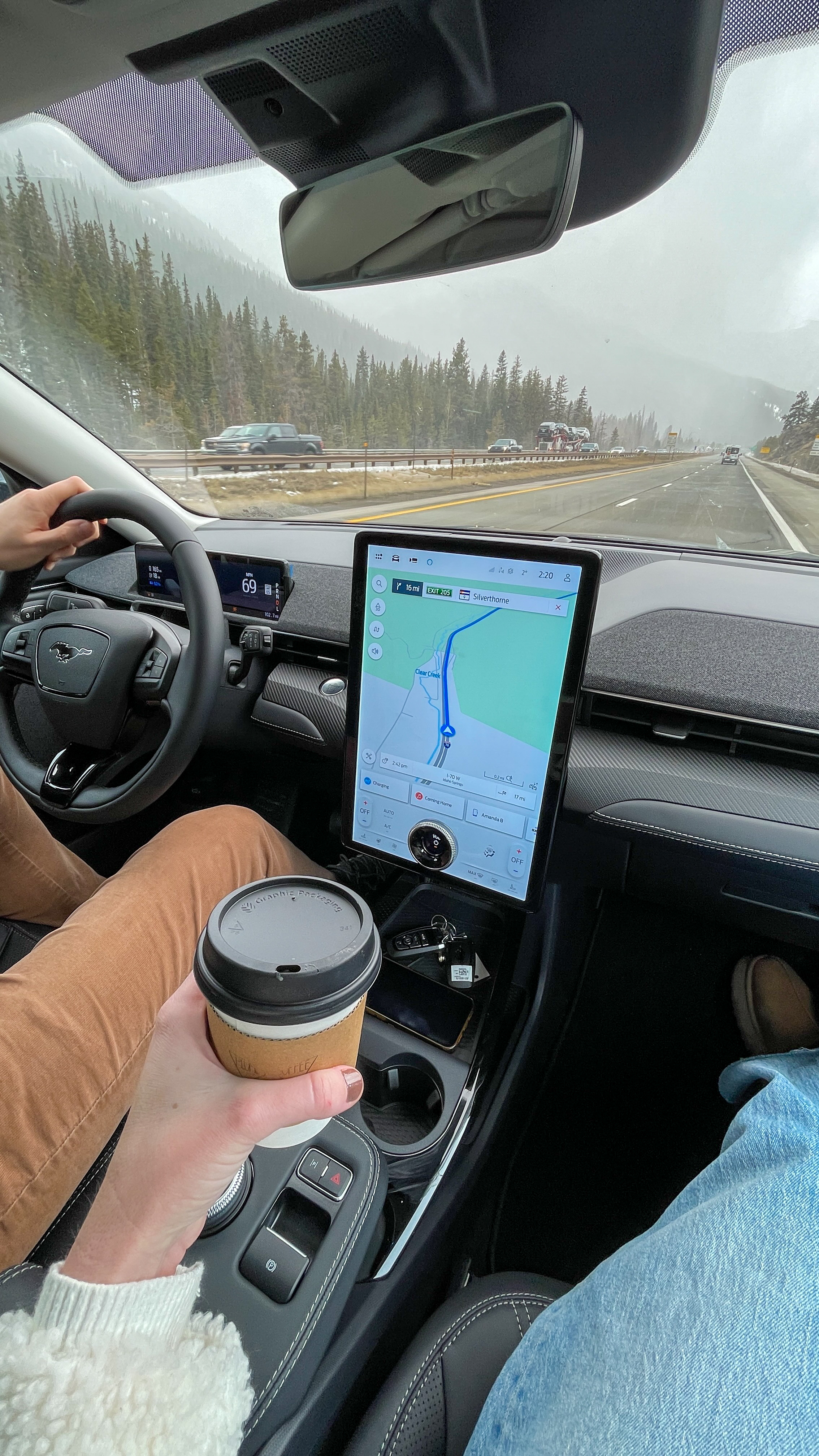 Amanda Bittner holds a coffee in the passenger seat as her travel partner drives the EV. The sky is overcast.