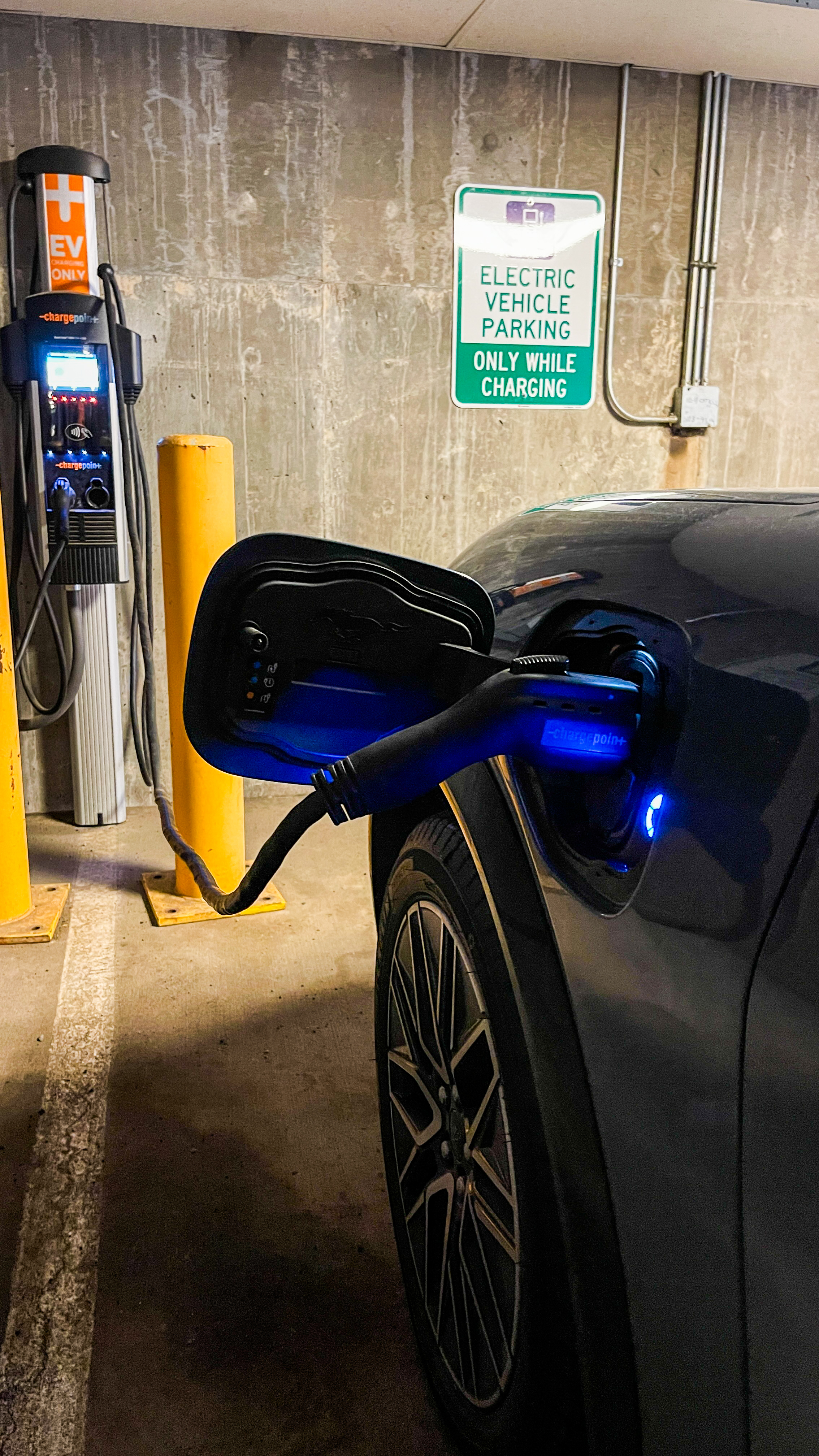 The Ford Mustang Mach-E is plugged in and charging in a covered parking garage. The charging port glows blue, creating a reflection on the car.