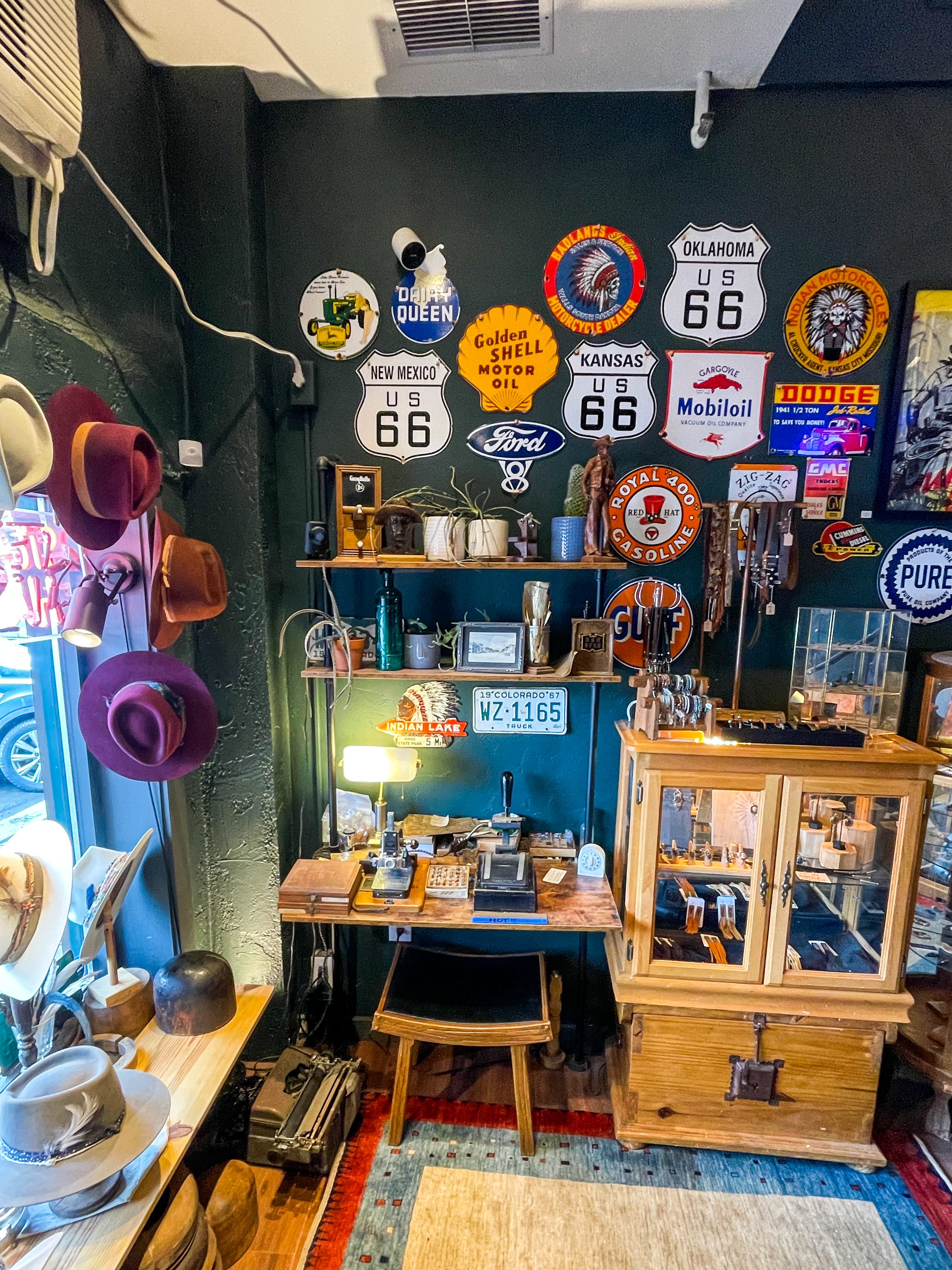 A craft table inside the hat maker’s shop. A collection of hats can be seen on the left, in a street-facing window, in multiple colors. On the deep green wall behind the craft table, numerous metal signs, including several of Route 66, can be seen.