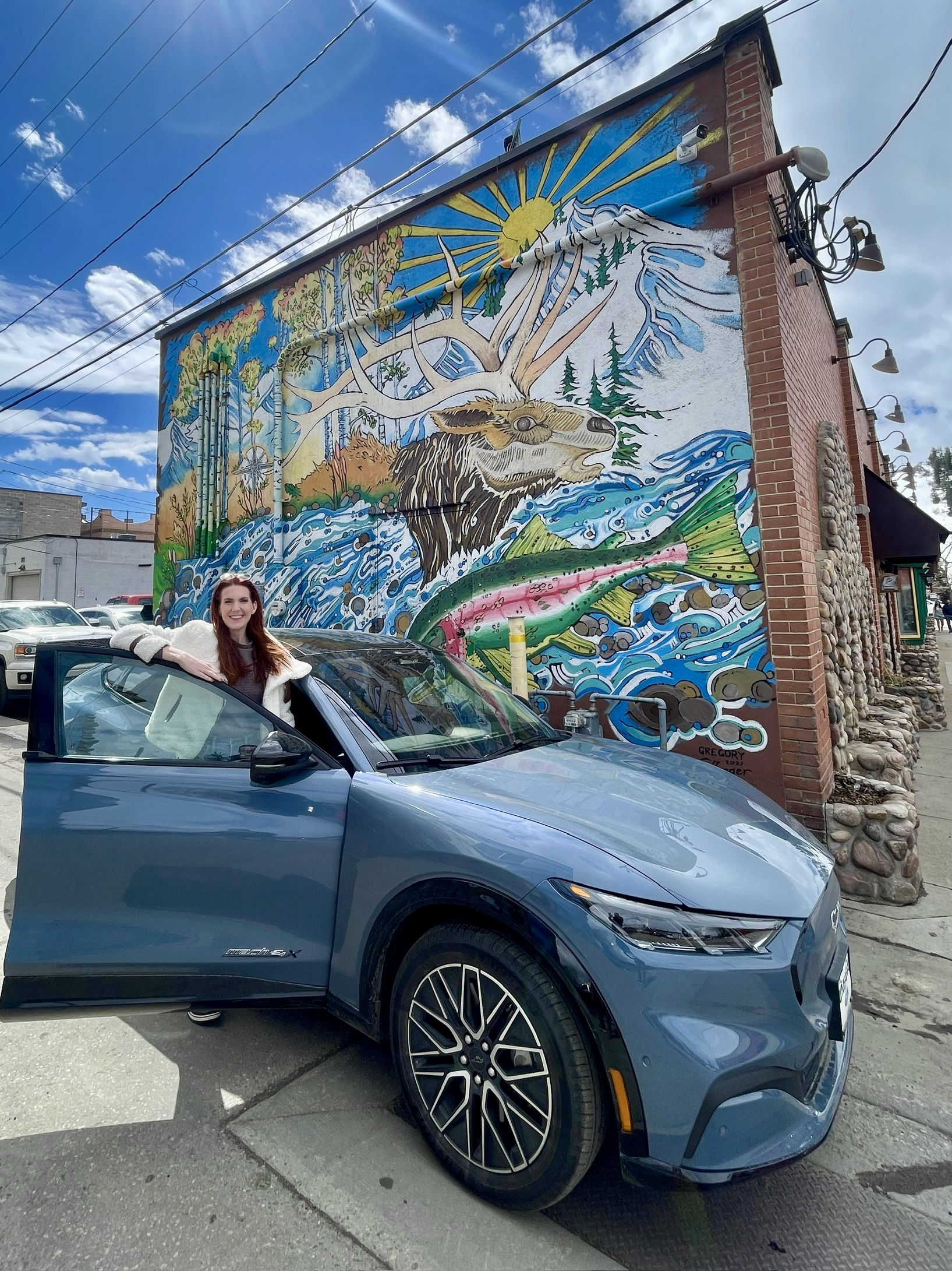 Amanda Bittner stands behind the open passenger door of her gray Ford Mustang Mach-E. The car is parked in front of a colorful mural painted on the side of a building and portrays the sun rising over a snow-capped mountain, with a moose and rainbow trout in the foreground.